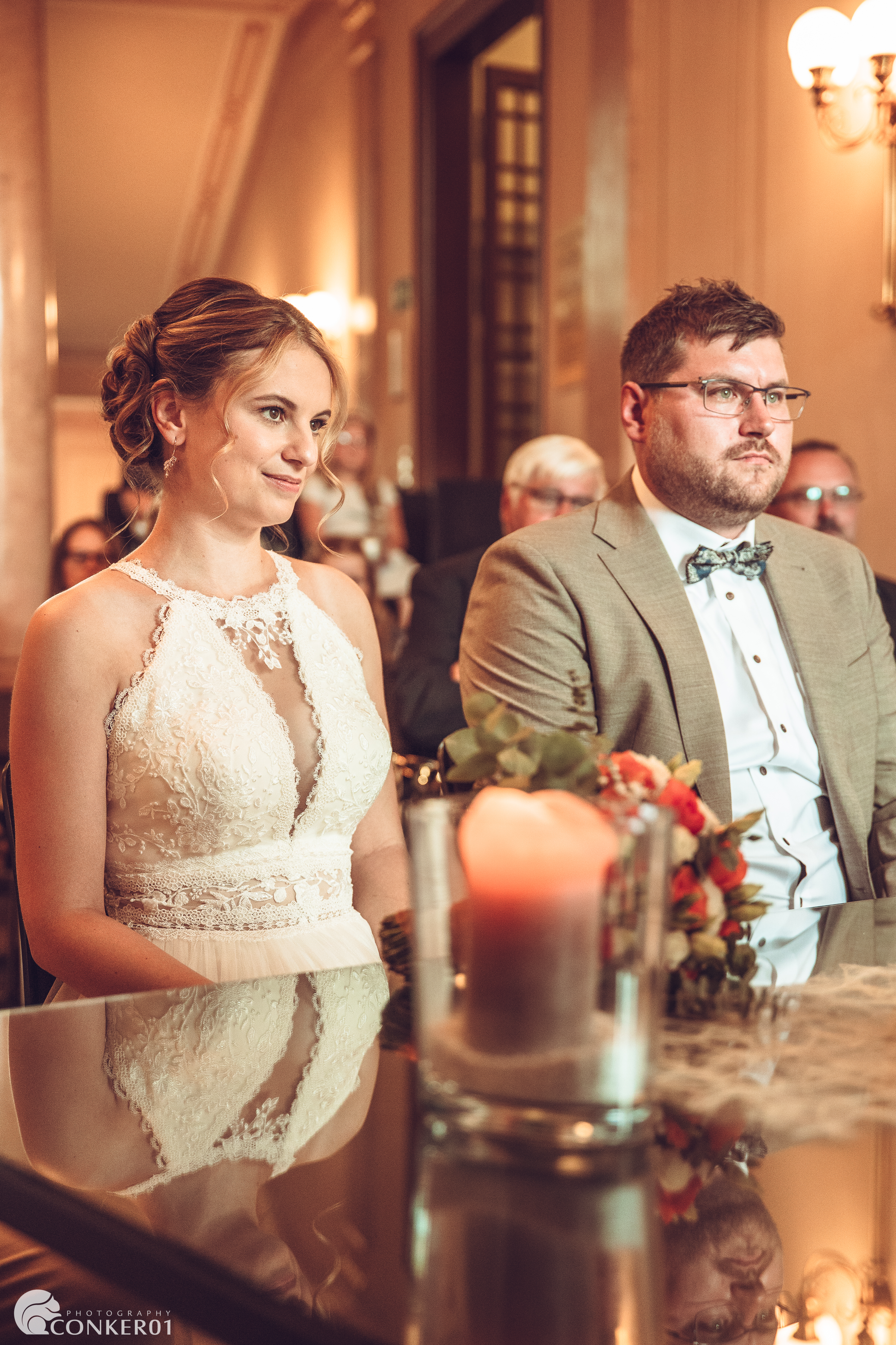 Brautpaar im Standesamt während der Rede der Standesbeamtin, Hochzeitsgesellschaft im Hintergrund.