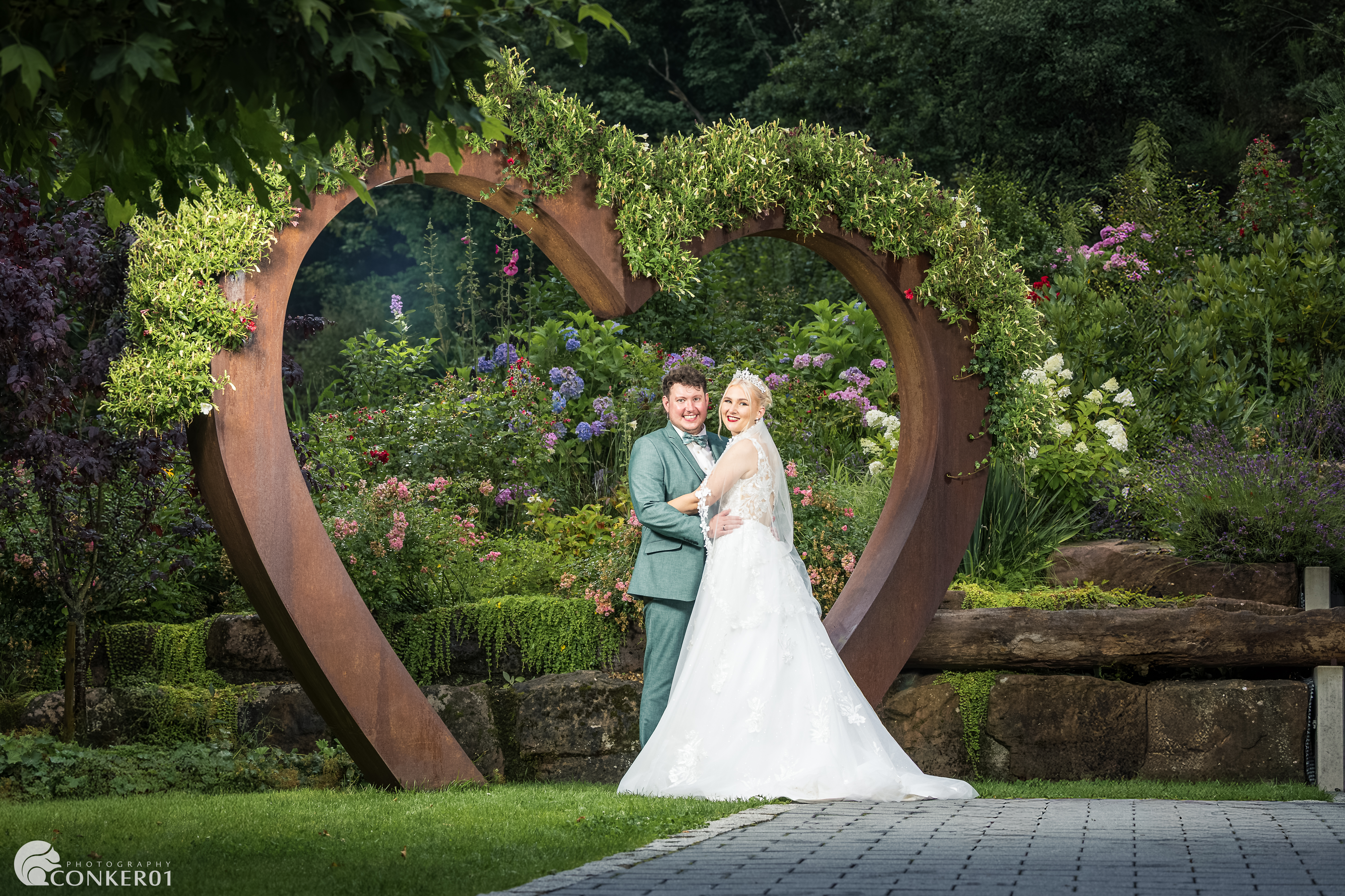 Lächelndes Brautpaar bei der Feier im Freien, stehend in einem mit Blumen bewachsenen Metallherz.