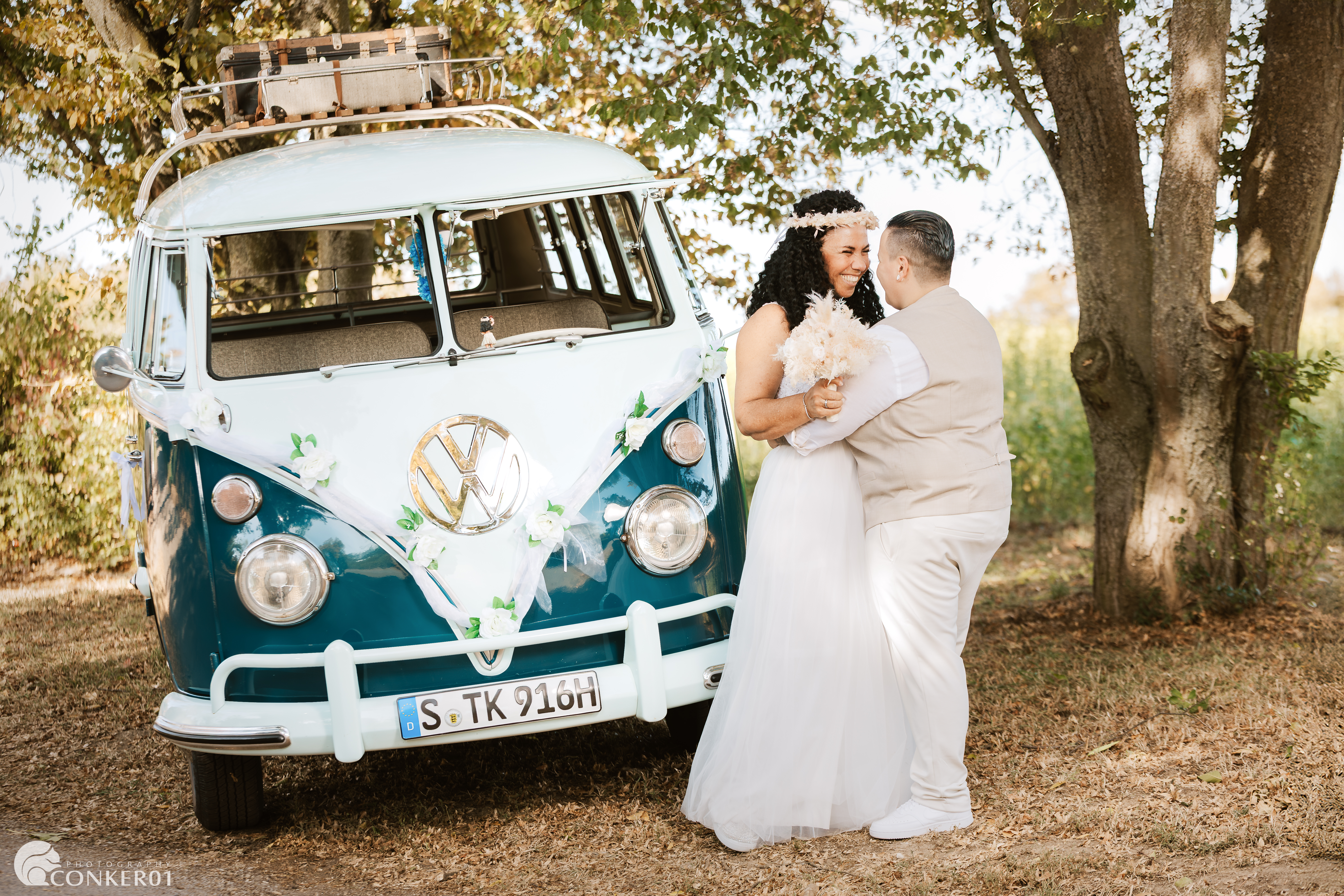 Zwei Bräute halten sich innig lächelnd vor einem alten VW-Bully als Hochzeitsauto.